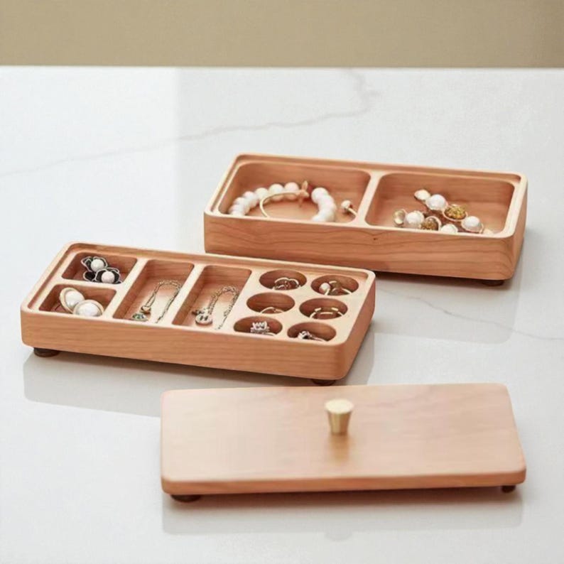 K&ouml;nnte beinhalten: Helle Holz-Schmuckorganisatoren mit mehreren F&auml;chern. Die Organizer enthalten verschiedene Schmuckst&uuml;cke, darunter Armb&auml;nder, Halsketten und Ringe. Ein passender Deckel mit einem goldfarbenen Knopf ist ebenfalls vorhanden.