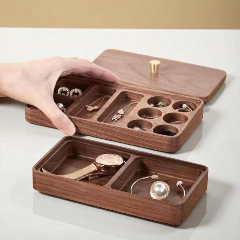 K&ouml;nnte beinhalten: Eine Holzschmuckschatulle mit Scharnierdeckel und mehreren F&auml;chern. Die Schatulle ist ge&ouml;ffnet und zeigt verschiedene Schmuckst&uuml;cke, darunter Ohrringe, Halsketten, Ringe, eine Uhr und ein Armband. Die Schatulle ist aus dunklem Holz gefertigt und hat einen goldfarbenen Griff.