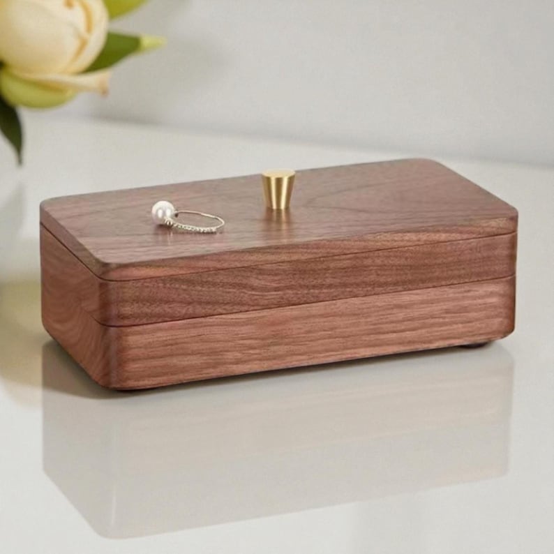 K&ouml;nnte beinhalten: Eine rechteckige Schmuckschatulle aus Holz mit einem Messingknopf oben. Die Schatulle hat eine warme braune Farbe und eine glatte, polierte Oberfl&auml;che. Ein silberner Ring mit einer Perle liegt oben auf der Schatulle. Die Schatulle ist zum Aufbewahren von Schmuck bestimmt.