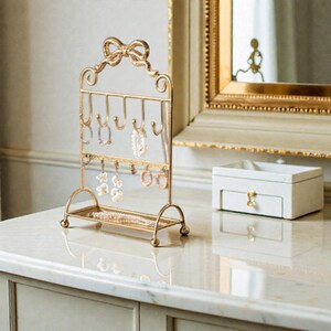 May include: A gold-colored metal jewelry organizer with a bow-shaped top. The organizer has hooks for hanging necklaces and earrings, and a small tray at the base. Several pieces of jewelry are displayed on the organizer.