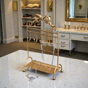 May include: A gold-colored metal jewelry organizer with a bow-shaped top. The organizer has hooks for necklaces and a small shelf. Two silver necklaces hang from the hooks. The organizer sits on a white marble surface.