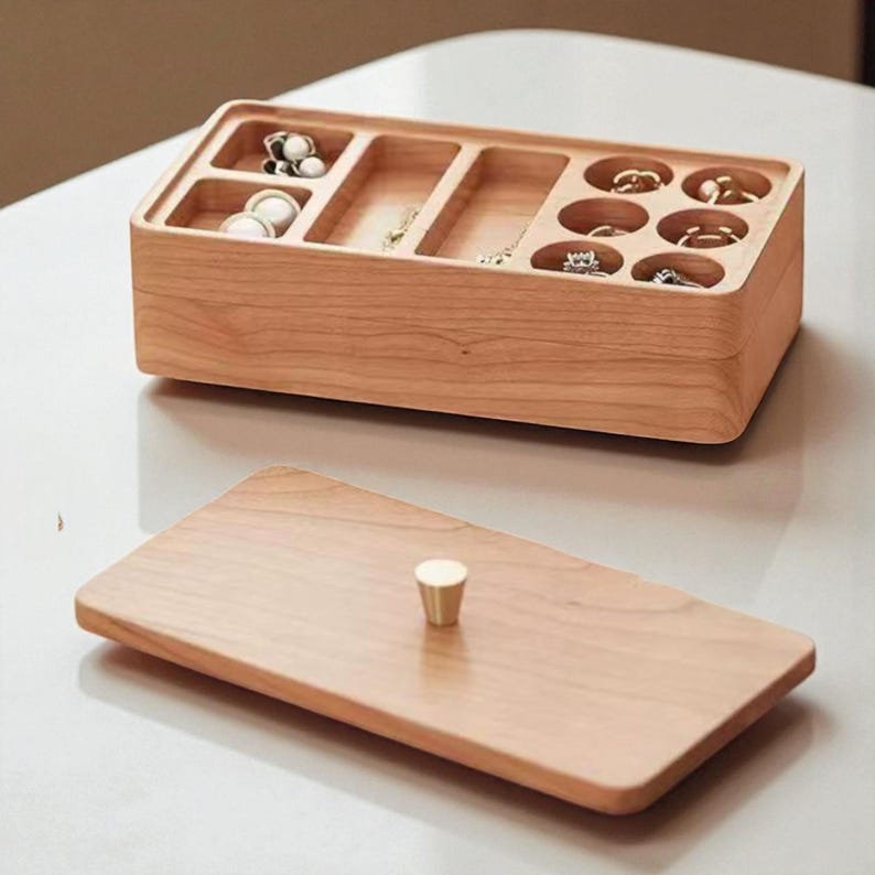 K&ouml;nnte beinhalten: Eine hellfarbene Schmuckschatulle aus Holz mit rechteckiger Form und mehreren F&auml;chern. Die Schatulle ist ge&ouml;ffnet und zeigt verschiedene Schmuckst&uuml;cke. Der Deckel ist separat, mit einem kleinen, goldfarbenen Knopf. Die Schatulle steht auf einer wei&szlig;en Oberfl&auml;che.