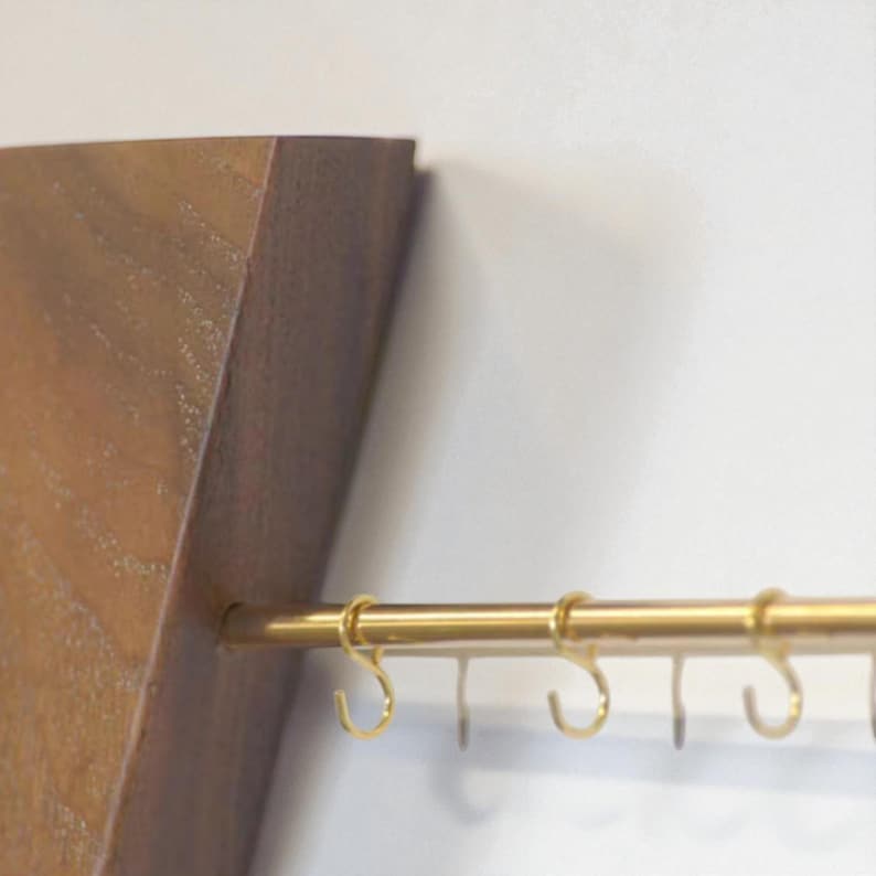May include: A wooden wall shelf with a brass rail and several small, gold-coloured hooks. The shelf is made of light brown wood and has a simple, modern design. The hooks are evenly spaced along the rail.