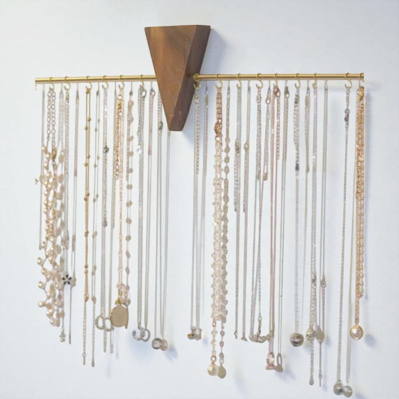 May include: A jewellery organiser made of wood and brass, displaying numerous necklaces. The design features a triangular wooden element at the top and a brass bar with hooks for the necklaces. The necklaces vary in style and length, creating a decorative display.