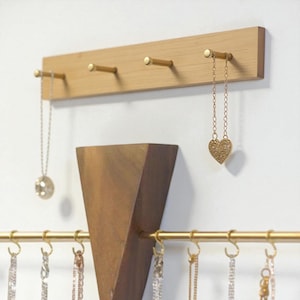 May include: A wooden wall-mounted jewelry organiser with four brass pegs. Two necklaces hang from the pegs. A gold-coloured heart pendant necklace and a silver-coloured round pendant necklace are displayed. A wooden geometric shelf is below.