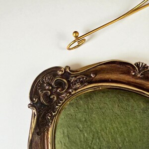 May include: Ornate, antique-style picture frame with a dark brown and gold finish. The frame has intricate detailing and a green velvet interior. A gold-colored, decorative hanging element is positioned above the frame.