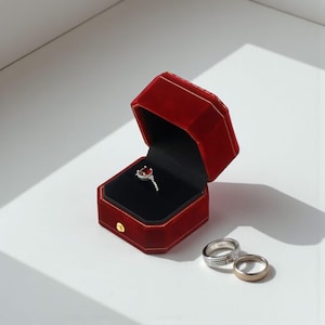 May include: A red velvet ring box with a silver ring with a red gemstone inside. Two additional rings are next to the box. The box is open, revealing a black interior. The box has a gold trim and a gold button.