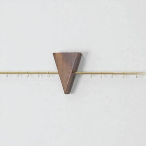 May include: A wall-mounted jewellery organiser featuring a triangular wooden accent piece and a brass rod with small hooks. The wooden piece is a dark brown colour, and the hooks are evenly spaced along the rod.