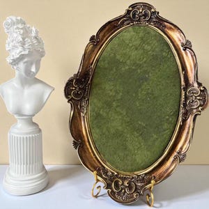 May include: An ornate, oval-shaped picture frame with a gold and brown finish, featuring a green interior. A white bust of a woman with curly hair is to the left. The frame is supported by a gold stand.