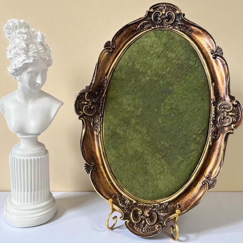 May include: An ornate, oval-shaped picture frame with a gold and brown finish, featuring a green interior. A white bust of a woman with curly hair is to the left. The frame is supported by a gold stand.
