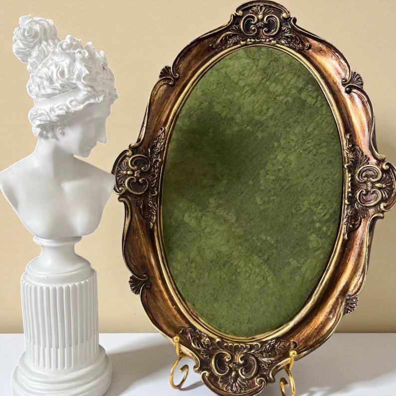 May include: An ornate, oval-shaped picture frame with a gold and brown finish, featuring a green interior. A white bust of a woman with detailed hair is positioned to the left of the frame. The frame is supported by a decorative stand.