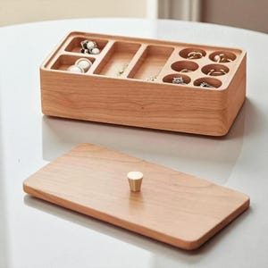 K&ouml;nnte beinhalten: Eine rechteckige Schmuckschatulle aus hellem Holz. Sie hat mehrere F&auml;cher f&uuml;r Ringe, Halsketten und Ohrringe. Der separate Deckel hat einen kleinen, goldfarbenen Knopf. Die Schatulle ist ge&ouml;ffnet und zeigt verschiedene Schmuckst&uuml;cke.