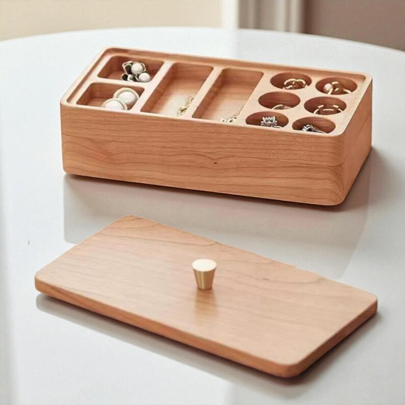 K&ouml;nnte beinhalten: Eine rechteckige Schmuckschatulle aus hellem Holz. Sie hat mehrere F&auml;cher f&uuml;r Ringe, Halsketten und Ohrringe. Der separate Deckel hat einen kleinen, goldfarbenen Knopf. Die Schatulle ist ge&ouml;ffnet und zeigt verschiedene Schmuckst&uuml;cke.