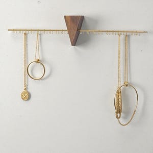 May include: A wall-mounted jewellery organiser with a triangular wooden base and a horizontal brass bar. Several gold necklaces with different pendants hang from the bar. The organiser is against a white wall, showcasing the jewellery.