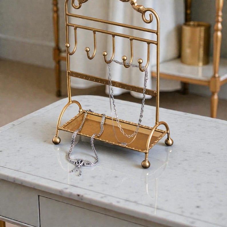 May include: A gold-colored metal jewelry stand with hooks and a tray. Several silver necklaces hang from the hooks and rest on the tray. The stand has a decorative, ornate design and sits on a white marble-topped table.