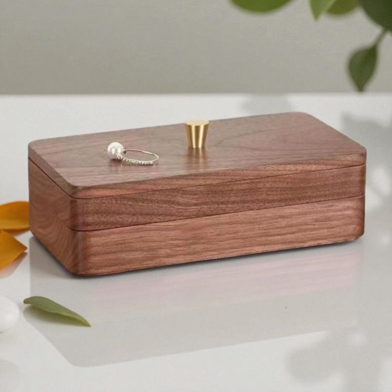 K&ouml;nnte beinhalten: Eine rechteckige Schmuckschatulle aus Holz mit einem Messingknopf oben. Ein silberner Ring mit einer Perle liegt auf der Schatulle. Die Schatulle ist aus dunkelbraunem Holz gefertigt und hat eine glatte Oberfl&auml;che. Die Schatulle ist geschlossen.