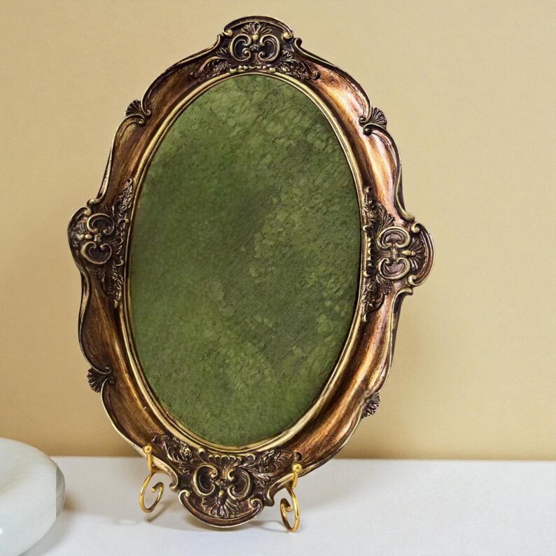 May include: Ornate, oval-shaped picture frame with a gold and bronze finish. The frame has decorative scrollwork and a green velvet backing. It stands on a gold-colored easel.