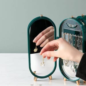 Peut inclure: Un organisateur de bijoux vert foncé avec un miroir et des accents dorés. L'organisateur a un miroir ovale et un compartiment pour ranger les bijoux. Une main tient une boucle d'oreille dorée devant le miroir.