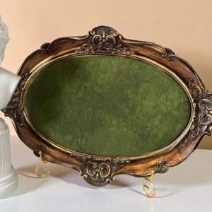 May include: An oval-shaped, ornate picture frame with a dark brown and gold finish. The frame has a green velvet backing and is supported by two gold-colored metal stands. The frame has decorative carvings.