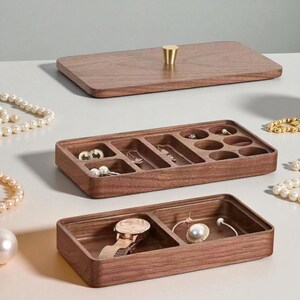 K&ouml;nnte beinhalten: Ein h&ouml;lzerner Schmuckorganizer mit rechteckiger Form und mehreren F&auml;chern. Der Organizer ist aus dunkelbraunem Holz gefertigt und hat einen passenden Deckel mit einem goldfarbenen Knopf. Die F&auml;cher enthalten verschiedene Schmuckst&uuml;cke, darunter Halsketten, Ohrringe und eine Uhr.