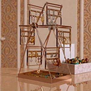 May include: A rose gold Ferris wheel-style jewellery organiser. It features multiple compartments for earrings and other jewellery. The base is a tray. The background shows a patterned wall and a box of decorative items.