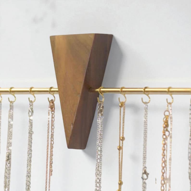 May include: A wooden jewellery organiser with a triangular design. The organiser is mounted on a white wall and features a gold-coloured bar with small hooks for hanging necklaces. Various necklaces in silver and gold tones are displayed.