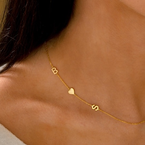 May include: A delicate gold necklace featuring the letters "B" and "S", separated by a small heart charm. The necklace is a fine chain, and the letters and heart are in a simple, elegant font. The necklace is worn on a person.