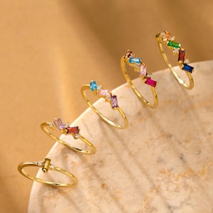 May include: Five gold rings with colorful baguette-cut gemstones are displayed. The rings feature a variety of colors, including blue, pink, green, and red. Each ring has a unique arrangement of stones, creating a vibrant and eye-catching design.