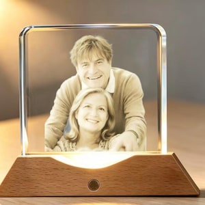 May include: A square, transparent acrylic photo frame featuring a sepia-toned portrait of two people. The frame is mounted on a light-coloured, trapezoidal wooden base with a circular power button and an integrated light source illuminating the image.