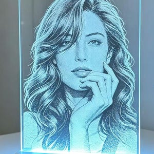 Può includere: Un pannello luminoso rettangolare in acrilico con un ritratto puntinato di una persona con lunghi capelli mossi, che appoggia il mento su una mano. Il pannello trasparente è illuminato da una luce blu fredda su una base in legno.