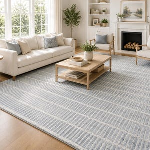 Op de afbeelding: Een groot rechthoekig vloerkleed met een herhalend patroon van lichtblauwe en witte verticale strepen met een getextureerde, licht verweerde afwerking. Het kleed bedekt een aanzienlijk deel van de vloer en toont een consistent lineair ontwerp.