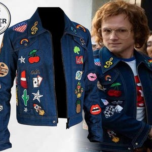 May include: A dark blue denim jacket featuring a variety of colourful embroidered patches including cherries, a rocket, a cactus, stars, lips, a pineapple, and musical notes. The jacket has a classic collar, long sleeves, and a front zip fastening.