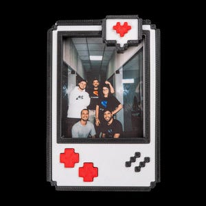 May include: A white and black pixelated picture frame in the shape of a retro handheld game console. The frame has red buttons and a red heart icon. The frame holds a color photograph of a group of people.