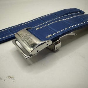 May include: A blue watch strap with white stitching and a silver clasp. The strap has a textured, crocodile-like pattern. The clasp is engraved with a logo. The strap is designed to be attached to a watch.