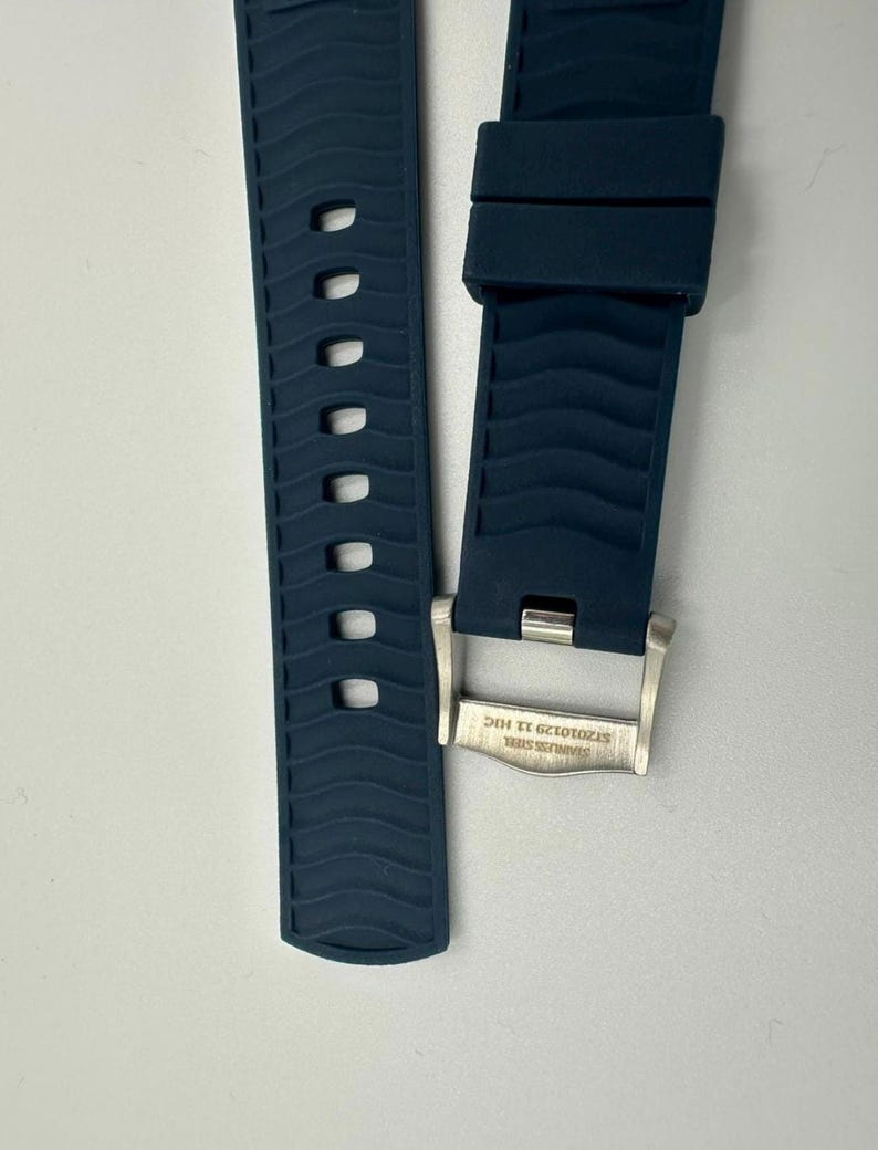 Puede incluir: Dos piezas de una correa de reloj azul oscuro con un patr&oacute;n de ondas texturizadas. Una pieza tiene una serie de agujeros ovalados. La otra pieza tiene una hebilla de acero inoxidable con el texto "STAINLESS STEEL ST2010129 11 HIC".