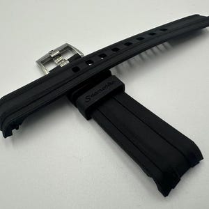 May include: Black rubber watch strap with a stainless steel buckle. The strap has a textured design and the word "Seamaster" is printed on the strap. The watch strap is lying on a white surface.