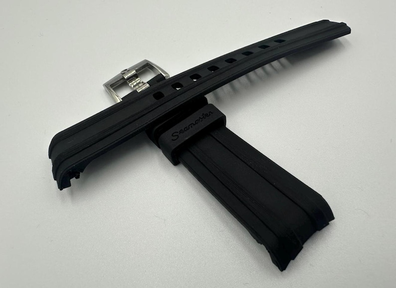 May include: Black rubber watch strap with a stainless steel buckle. The strap has a textured design and the word "Seamaster" is printed on the strap. The watch strap is lying on a white surface.