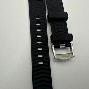 May include: Black watch band with a textured wave pattern and a stainless steel buckle. The band has a series of oval cutouts. The buckle has engraved text.