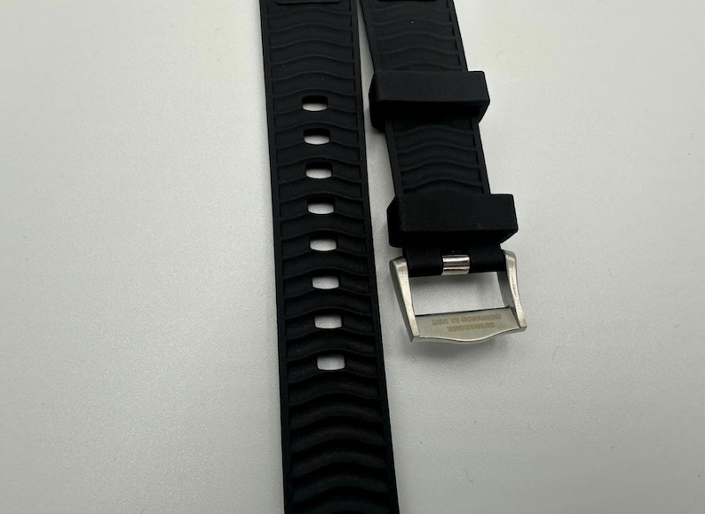 May include: Black watch band with a textured wave pattern and a stainless steel buckle. The band has a series of oval cutouts. The buckle has engraved text.