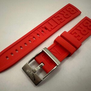 May include: A vibrant red watch strap featuring the embossed brand name "BREITLING". The strap is paired with a silver-coloured buckle. The strap is made of a flexible material and is designed for a watch.