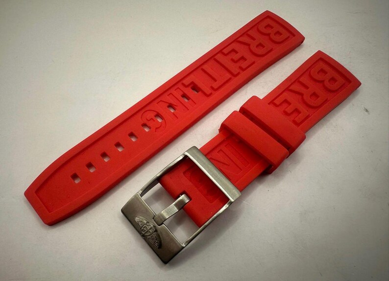 May include: A vibrant red watch strap featuring the embossed brand name "BREITLING". The strap is paired with a silver-coloured buckle. The strap is made of a flexible material and is designed for a watch.