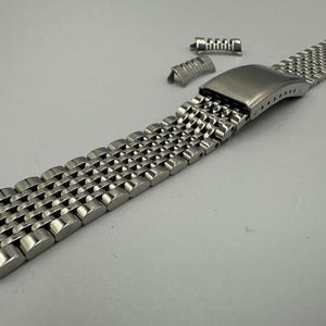 May include: A stainless steel watch band with a unique, textured design. The band is composed of small, interconnected links, creating a flexible and durable strap. Additional pieces are included for adjustment or replacement. The band is designed to attach to a watch case.