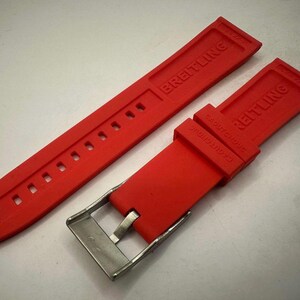 May include: A vibrant red watch strap, featuring the embossed brand name "BREITLING". The strap includes a stainless steel buckle and several adjustment holes. This watch strap is designed for a Breitling watch.