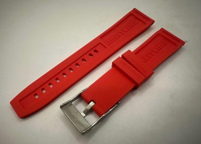 May include: A vibrant red watch strap, featuring the embossed brand name "BREITLING". The strap includes a stainless steel buckle and several adjustment holes. This watch strap is designed for a Breitling watch.