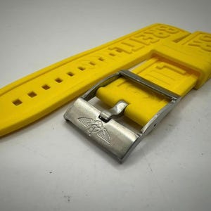 Puede incluir: Correa de reloj amarilla brillante con hebilla plateada. La correa tiene el nombre de la marca en relieve en may&uacute;sculas. La hebilla presenta un emblema alado. La correa es de material flexible.