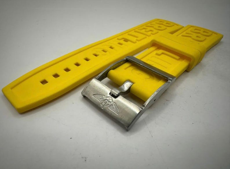 Puede incluir: Correa de reloj amarilla brillante con hebilla plateada. La correa tiene el nombre de la marca en relieve en may&uacute;sculas. La hebilla presenta un emblema alado. La correa es de material flexible.