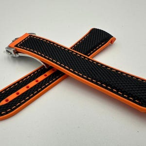 May include: Two-piece watch strap in black with orange accents. The strap features a textured black surface with orange edges and stitching. The strap has a stainless steel clasp.