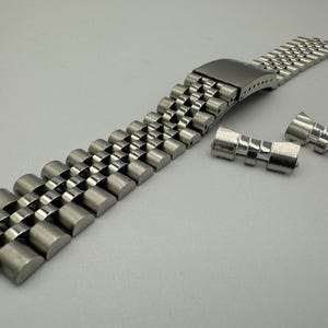 May include: A silver-coloured stainless steel watch strap with a classic design. The strap is made up of numerous small, rounded links, and includes a clasp and two additional connecting pieces. The watch strap appears to be new.
