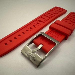 May include: Bright red watch strap with the brand name "BREITLING" embossed. The strap features a silver-coloured buckle with the brand's logo. The strap is made of a flexible material and has several holes for adjustment.