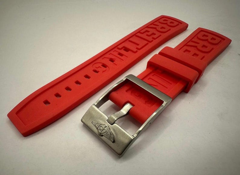 May include: Bright red watch strap with the brand name "BREITLING" embossed. The strap features a silver-coloured buckle with the brand's logo. The strap is made of a flexible material and has several holes for adjustment.